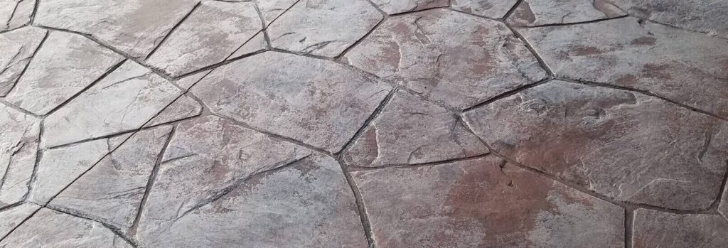 Pavimento de hormigón impreso con acabado de piedra irregular y manchas blanquecinas distribuidas por la superficie.